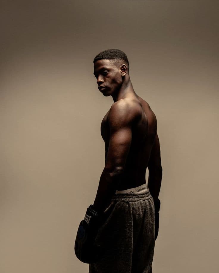 A muscular young man, bare-chested, wearing a black boxing glove and looking over his shoulder