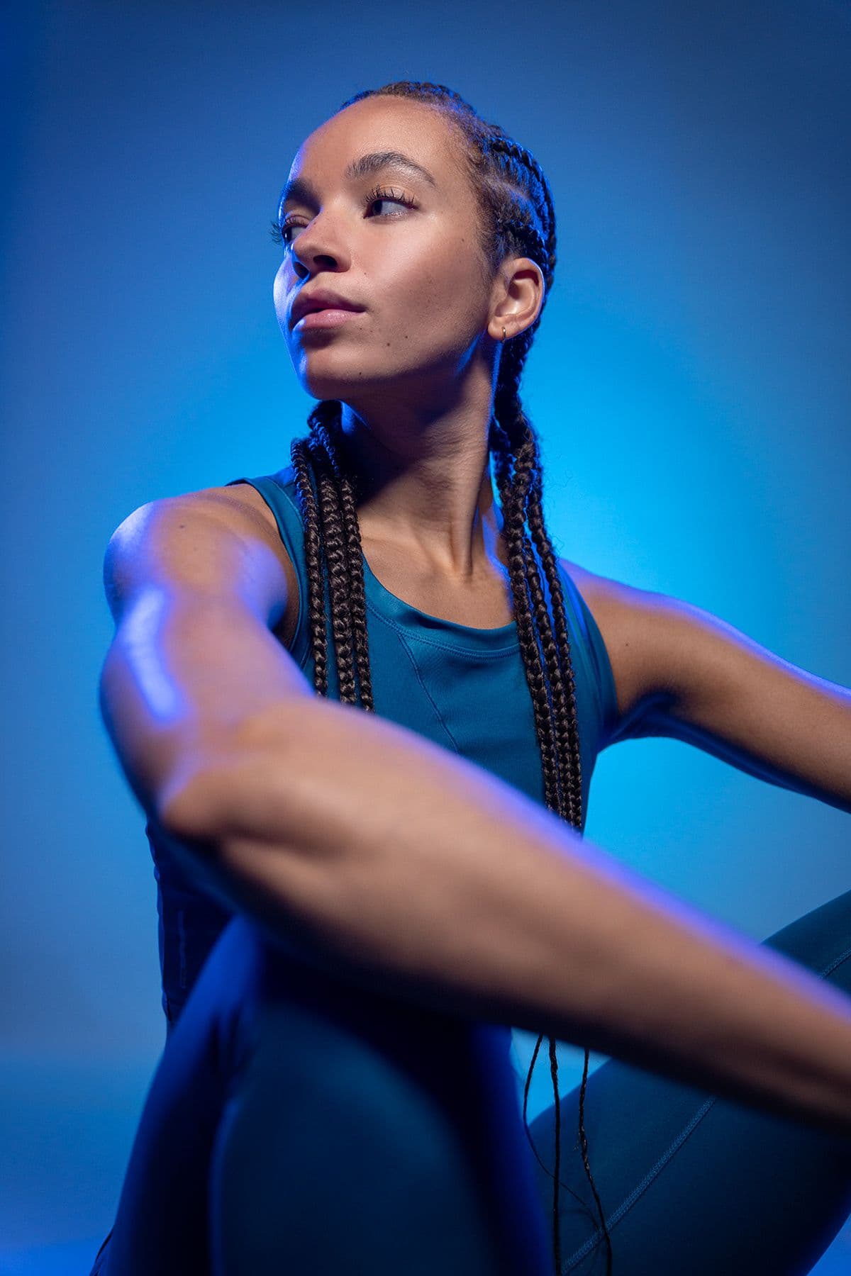 A young woman with long braids in a teal tracksuit stretching under blue light